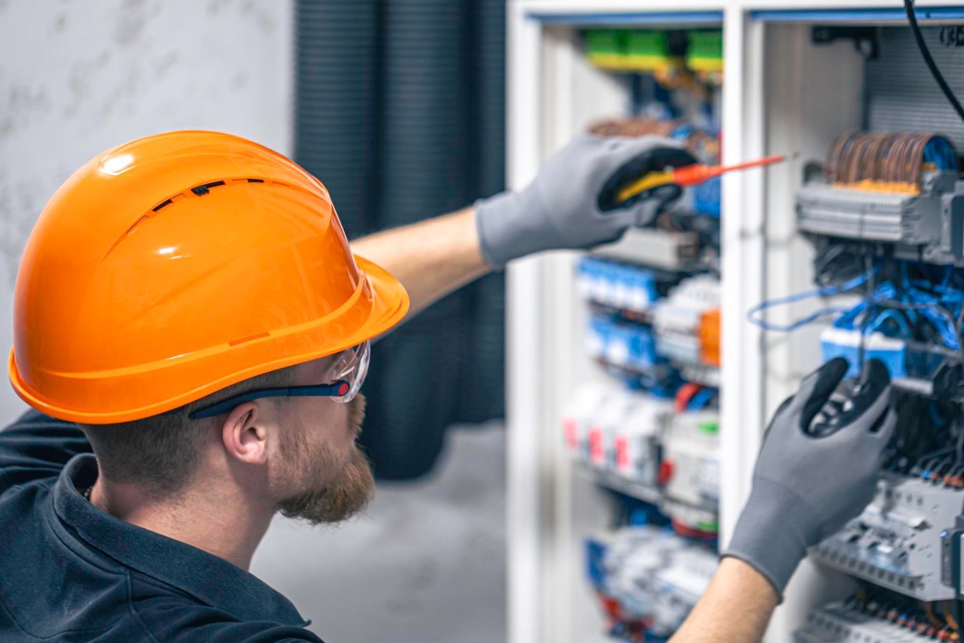 Electrician working on a circuit panel, wearing safety glasses, gloves, and an orange hard hat.