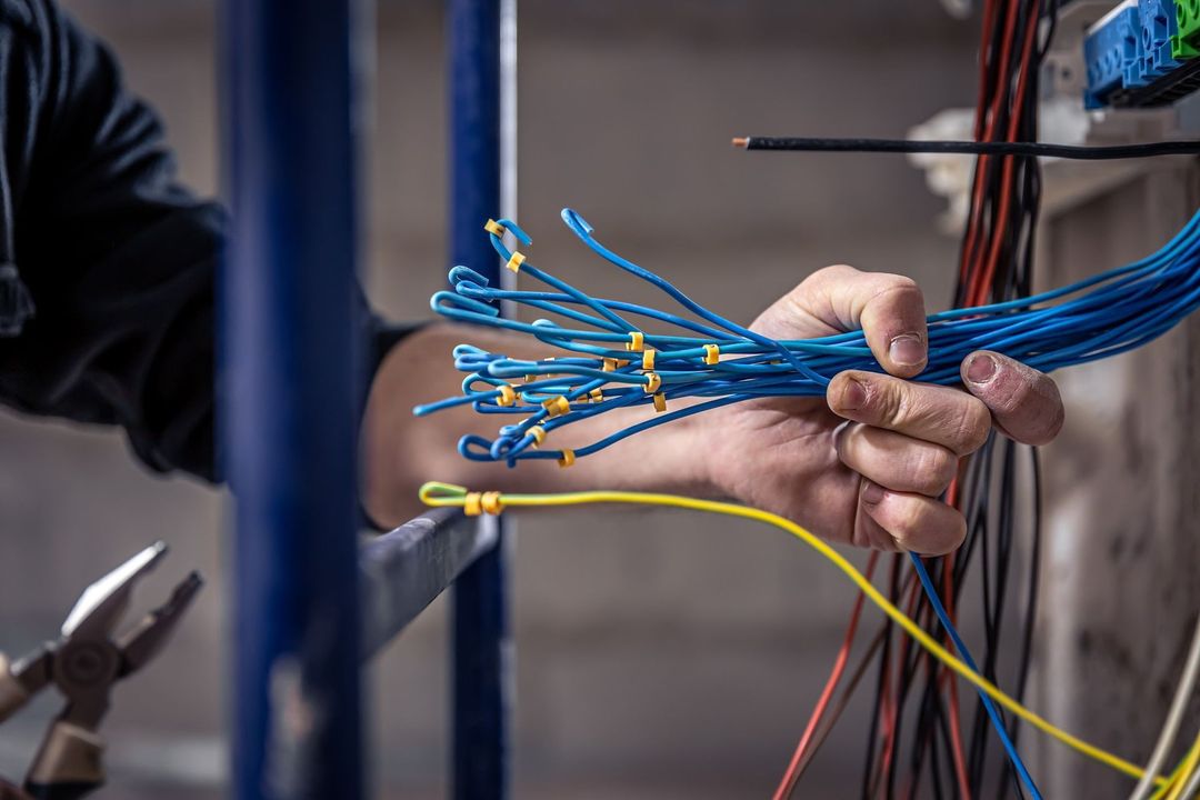 Person's hand holding blue electrical wires, working with other wires and tools on a construction site.