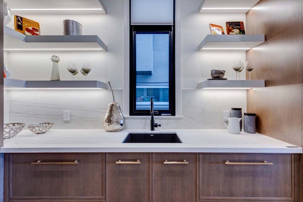 Modern kitchen with white countertop, dark sink, and floating shelves with lit undersides, and black window.