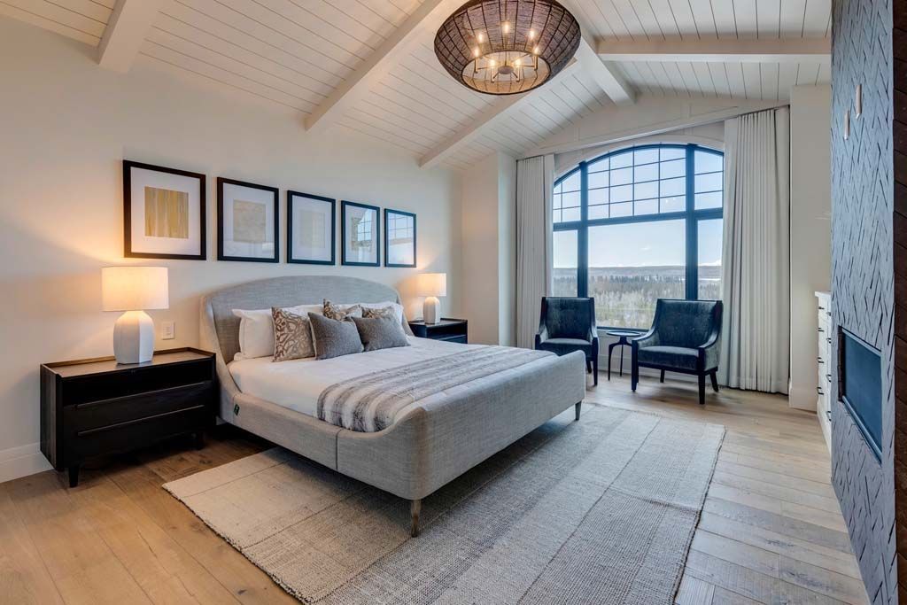 Bedroom with arched window, bed, and chairs. White walls, wooden floor, decorative chandelier.
