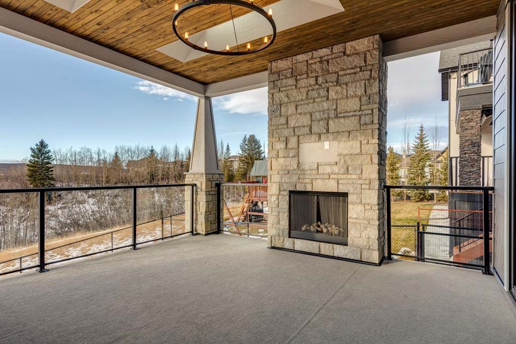Covered outdoor patio with a stone fireplace, glass railing, and a view of trees and a bright blue sky.