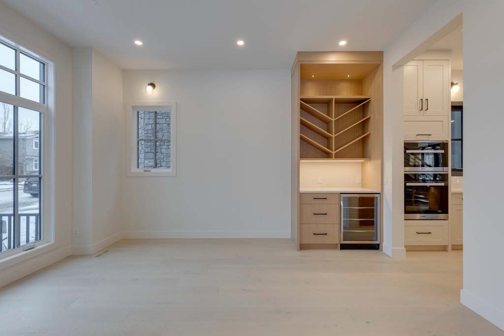 Empty, light-filled room with wood flooring and built-in wine storage, adjacent to a kitchen area with appliances.
