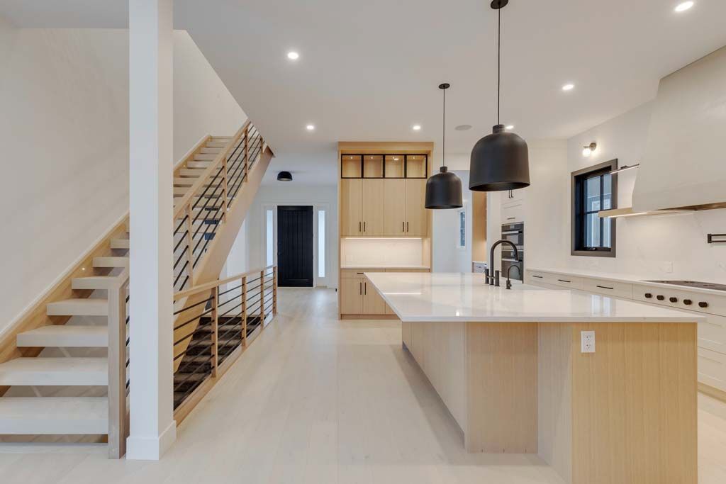 Modern kitchen with light wood cabinetry and island, black pendant lights, and staircase.