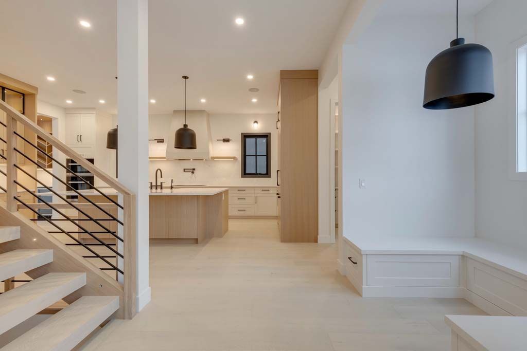 Bright, modern home interior with staircase and open kitchen. Light wood flooring and cabinetry, black light fixtures.