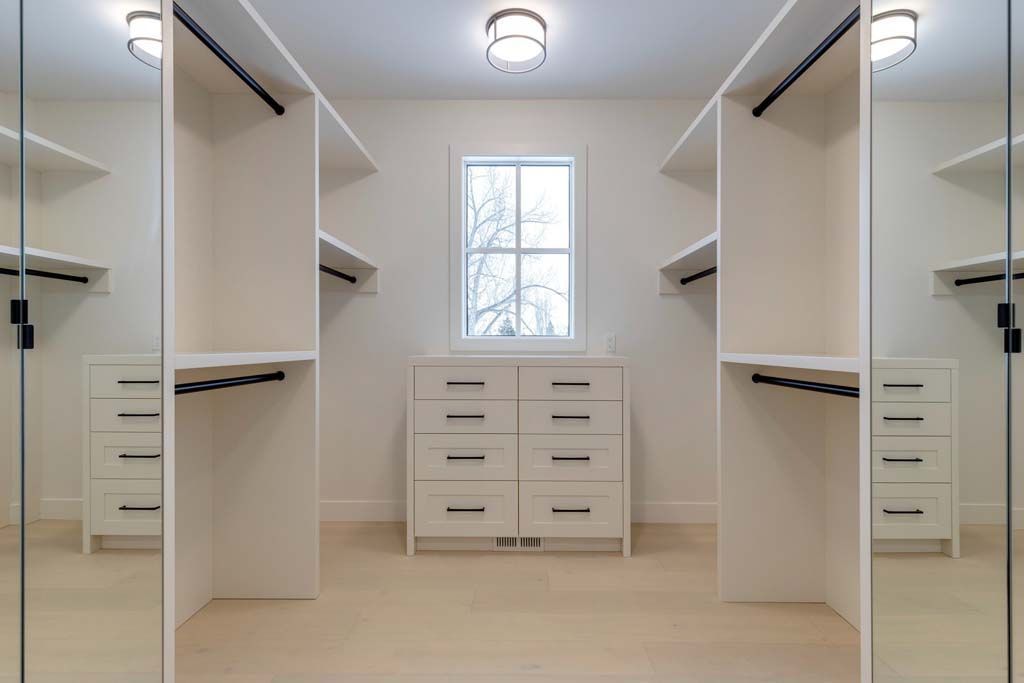 Walk-in closet with white shelves, drawers, and full-length mirrors, with a window centered at the back.