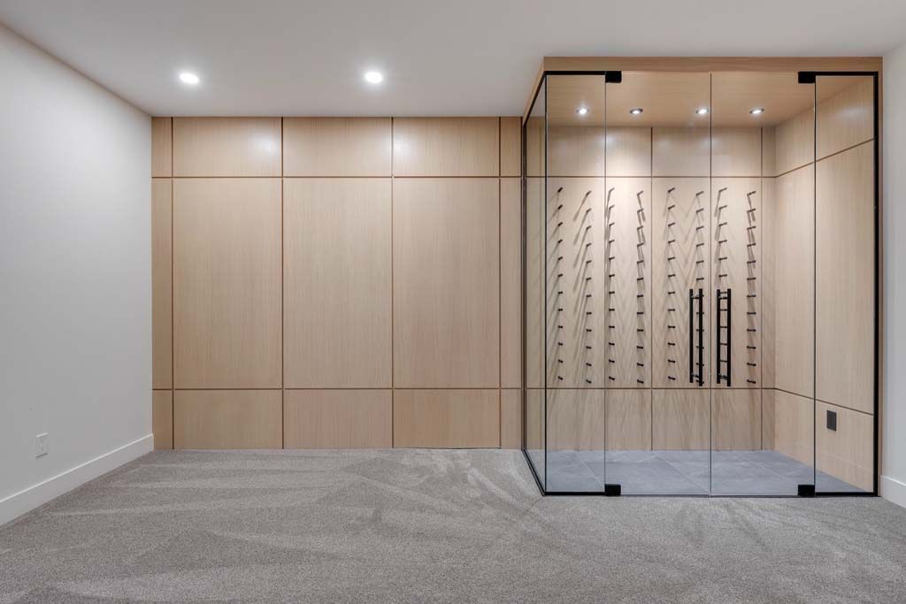 Empty room with a glass-enclosed wine cellar and light wood paneling. Gray carpet.