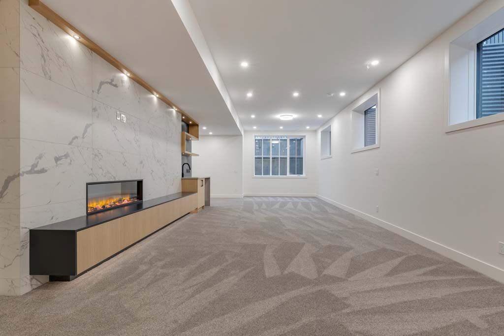 Empty, newly-renovated basement room with a fireplace, marble wall, carpet, and windows.