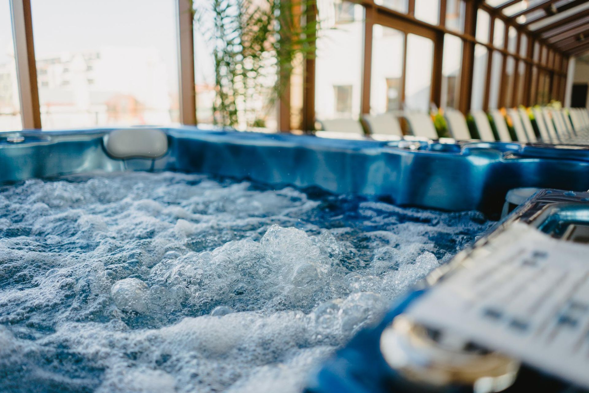 Bubbling blue hot tub in a sunlit room, with windows and lounge chairs visible in the background.