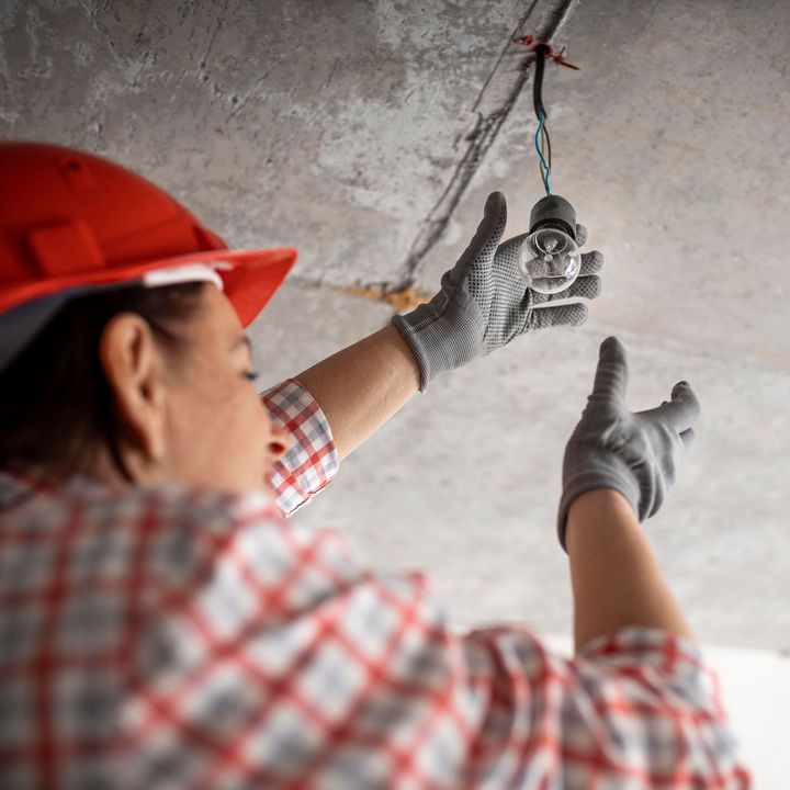 Person in red hard hat and gloves working on exposed wiring on a concrete ceiling.