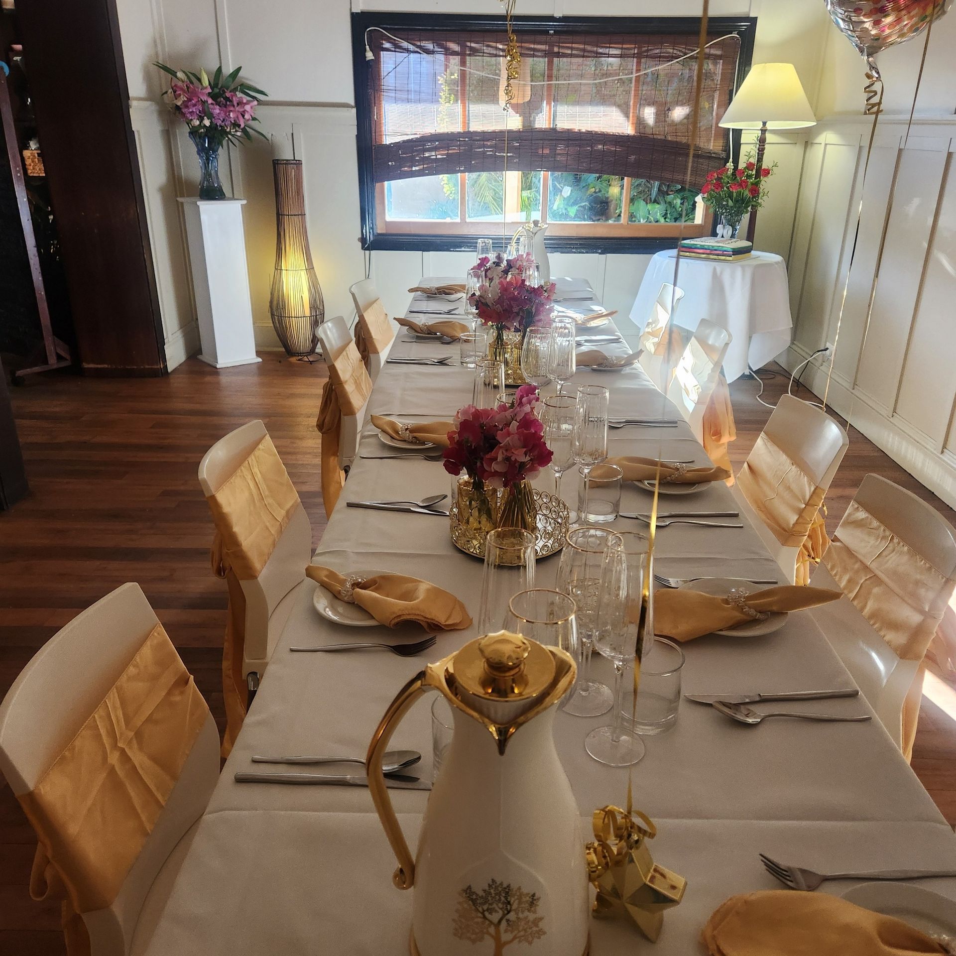 Long table set for a formal event with gold and white accents, floral centerpieces, and a window in the background.