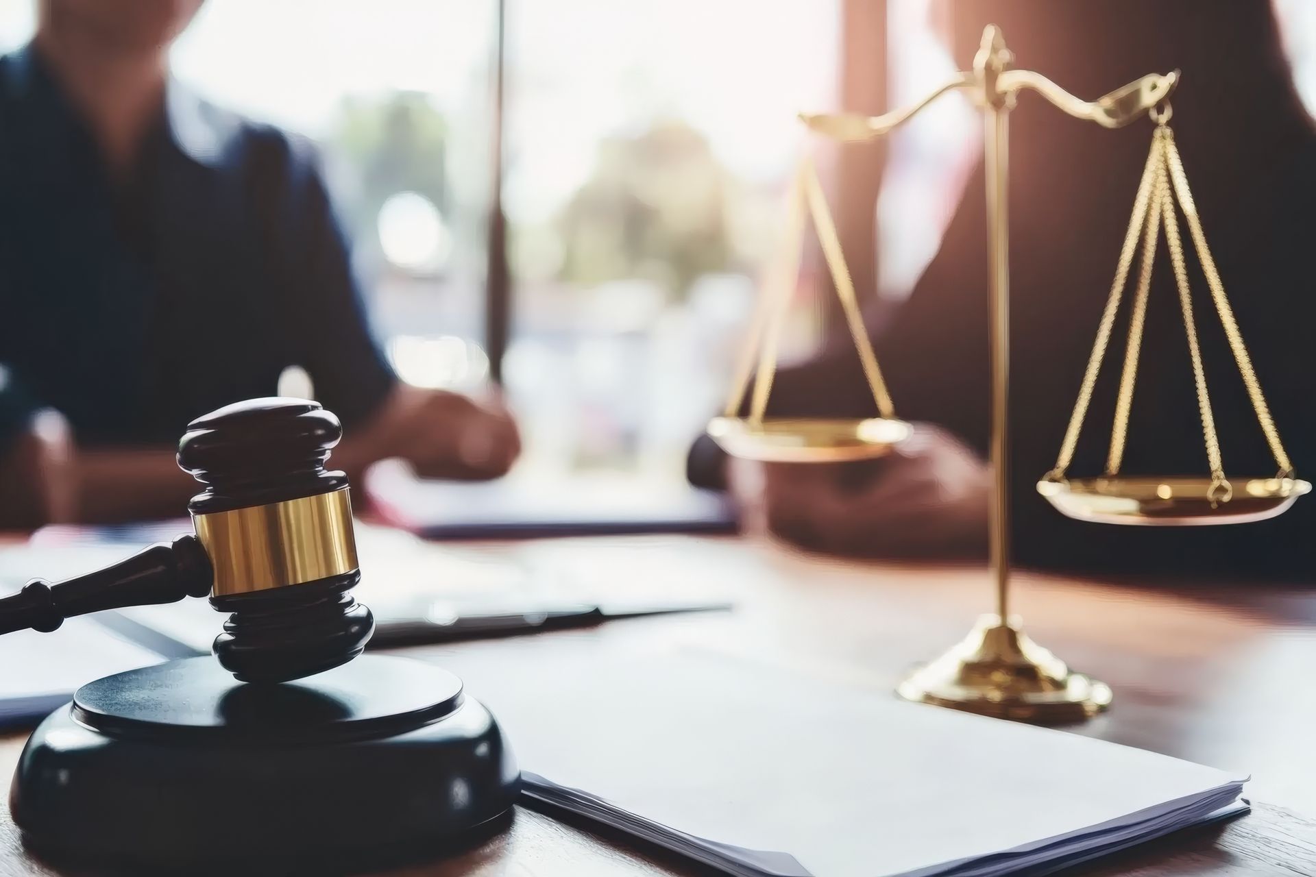 A wooden gavel and gold-colored scales of justice sit on a desk in a law office with people out of focus in the background.