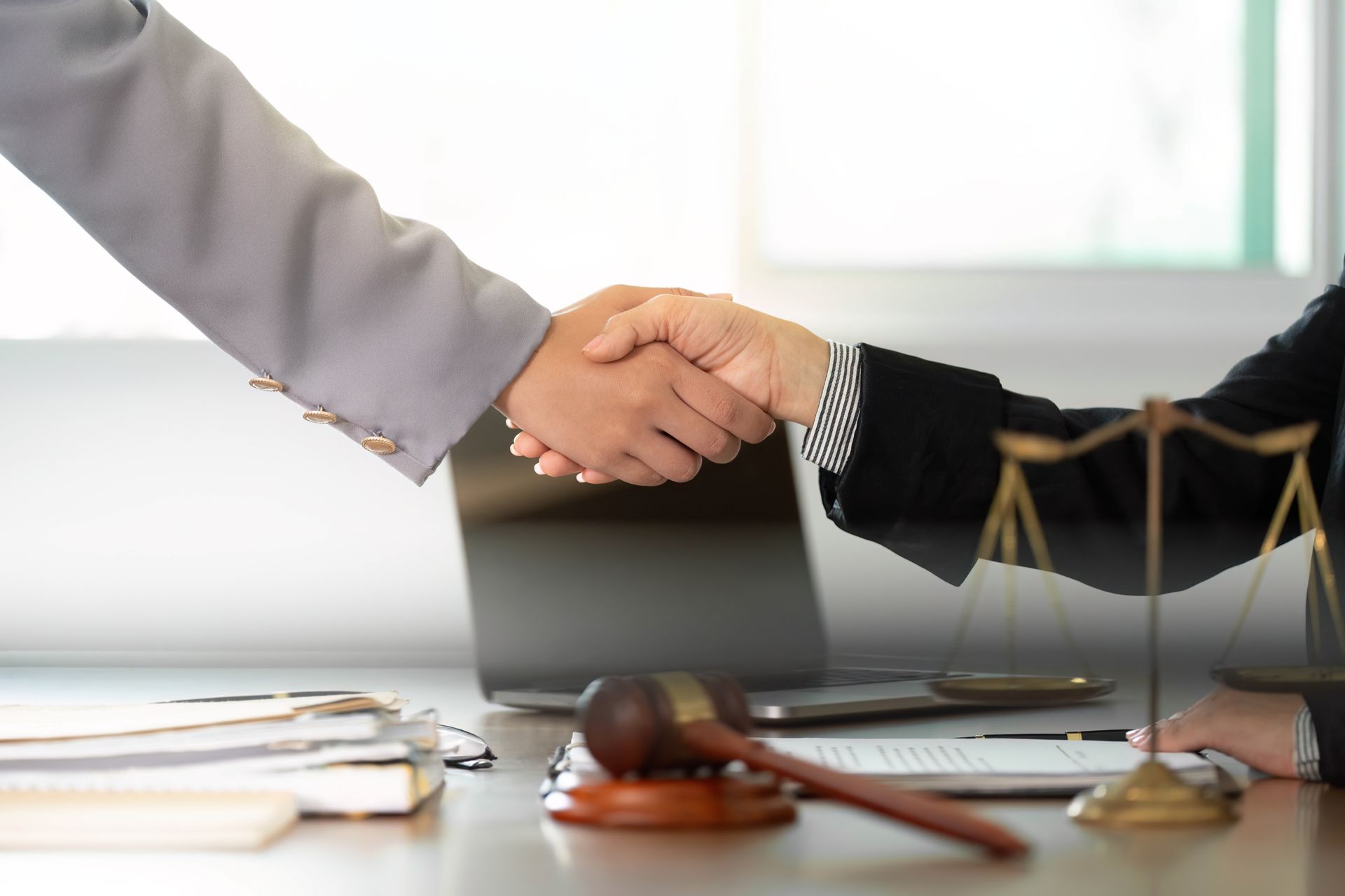 Two people in business attire shake hands over a desk with a gavel, legal scales, and documents.