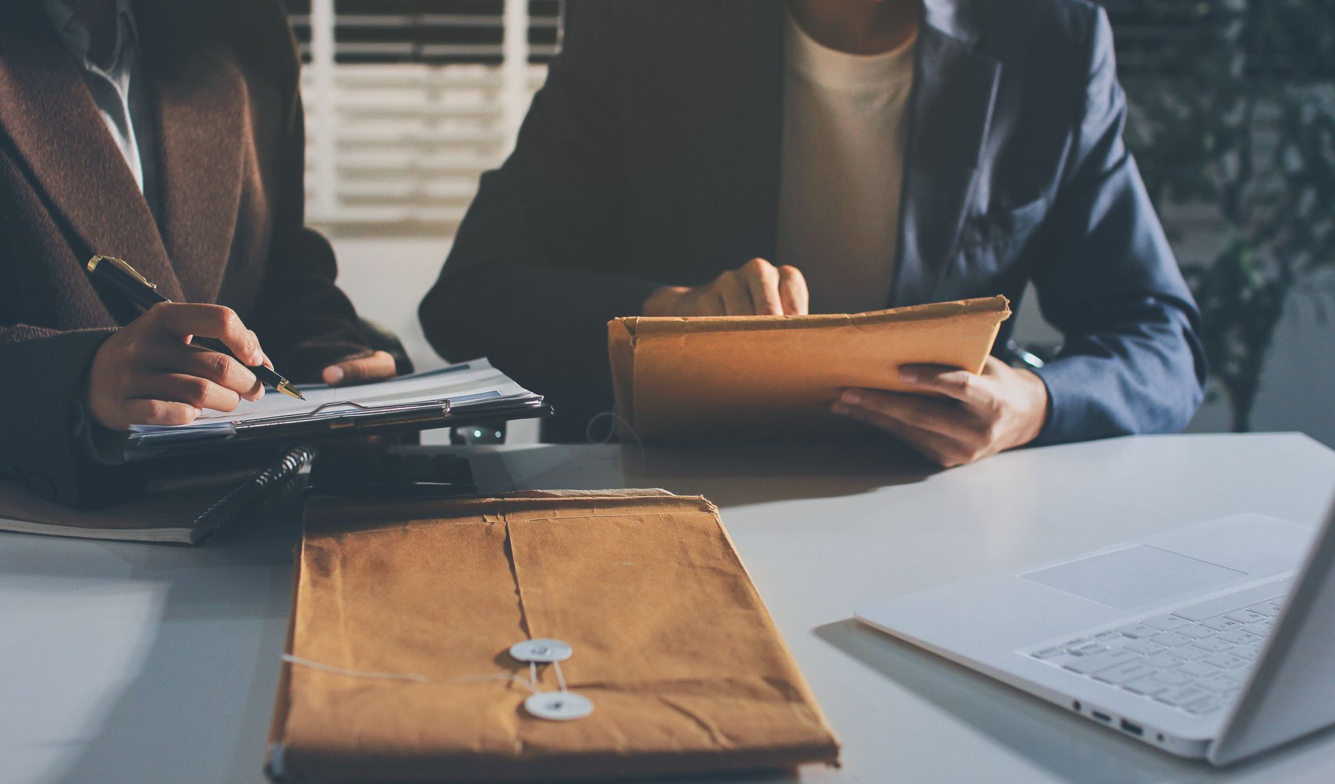 Two people sit at a desk reviewing documents and folders in an office setting.