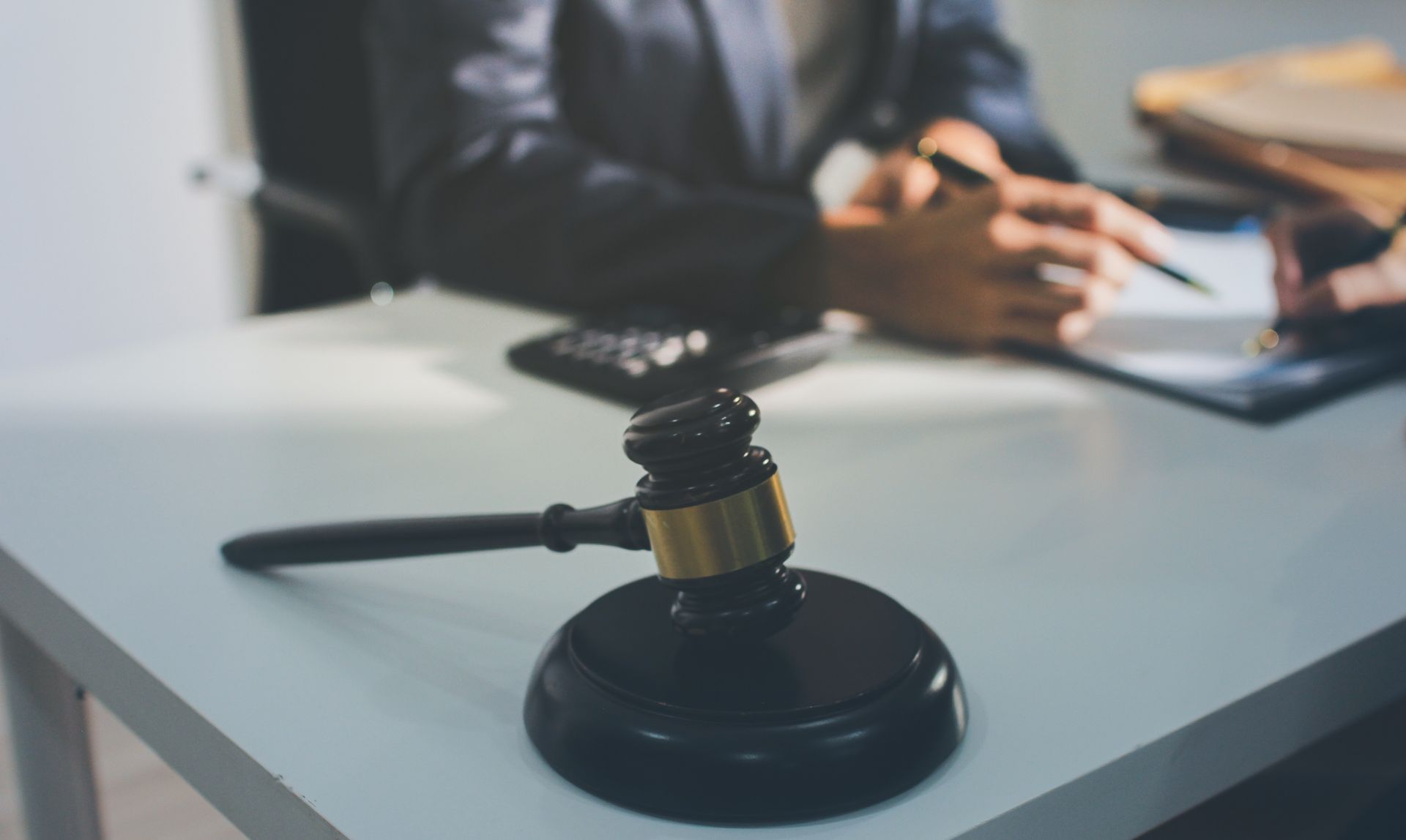 Gavel on a desk in front of a person in a suit, blurred. A calculator and papers are nearby.