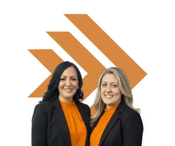 Two women in orange tops and black jackets smile in front of an orange logo.