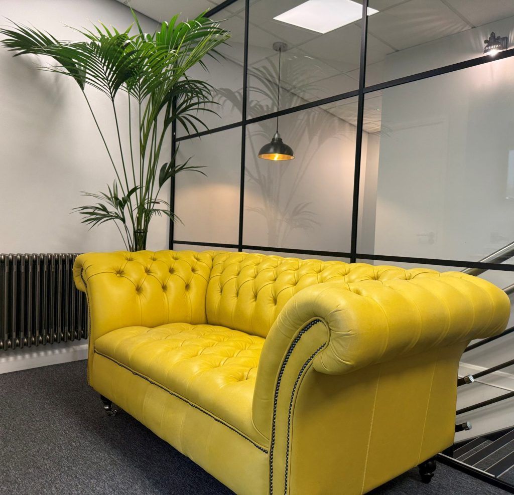 Yellow tufted sofa in a modern office lounge with glass walls, a hanging lamp, and a tall potted palm.