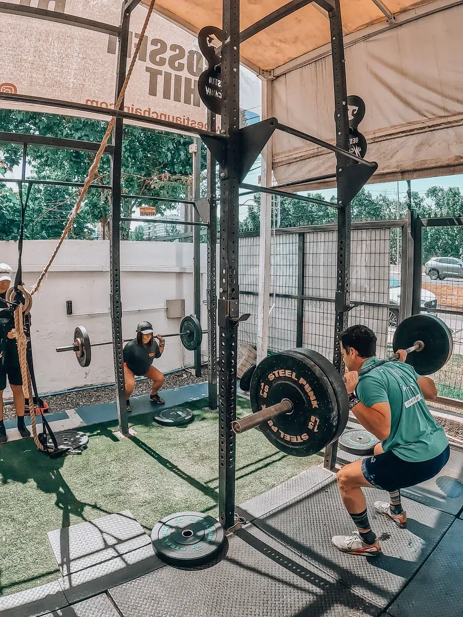 Gente haciendo sentadillas con pesas en un gimnasio al aire libre. Racks de sentadillas negros, césped verde y camisetas azules.