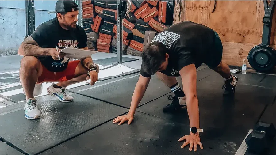Hombre en posición de ejercicio, supervisado por un entrenador en un gimnasio. Ambos visten ropa deportiva.