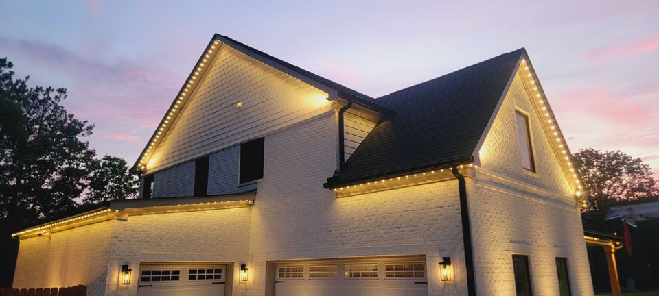 Two-story house with warm string lights along the roof at dusk under a pink-blue sky