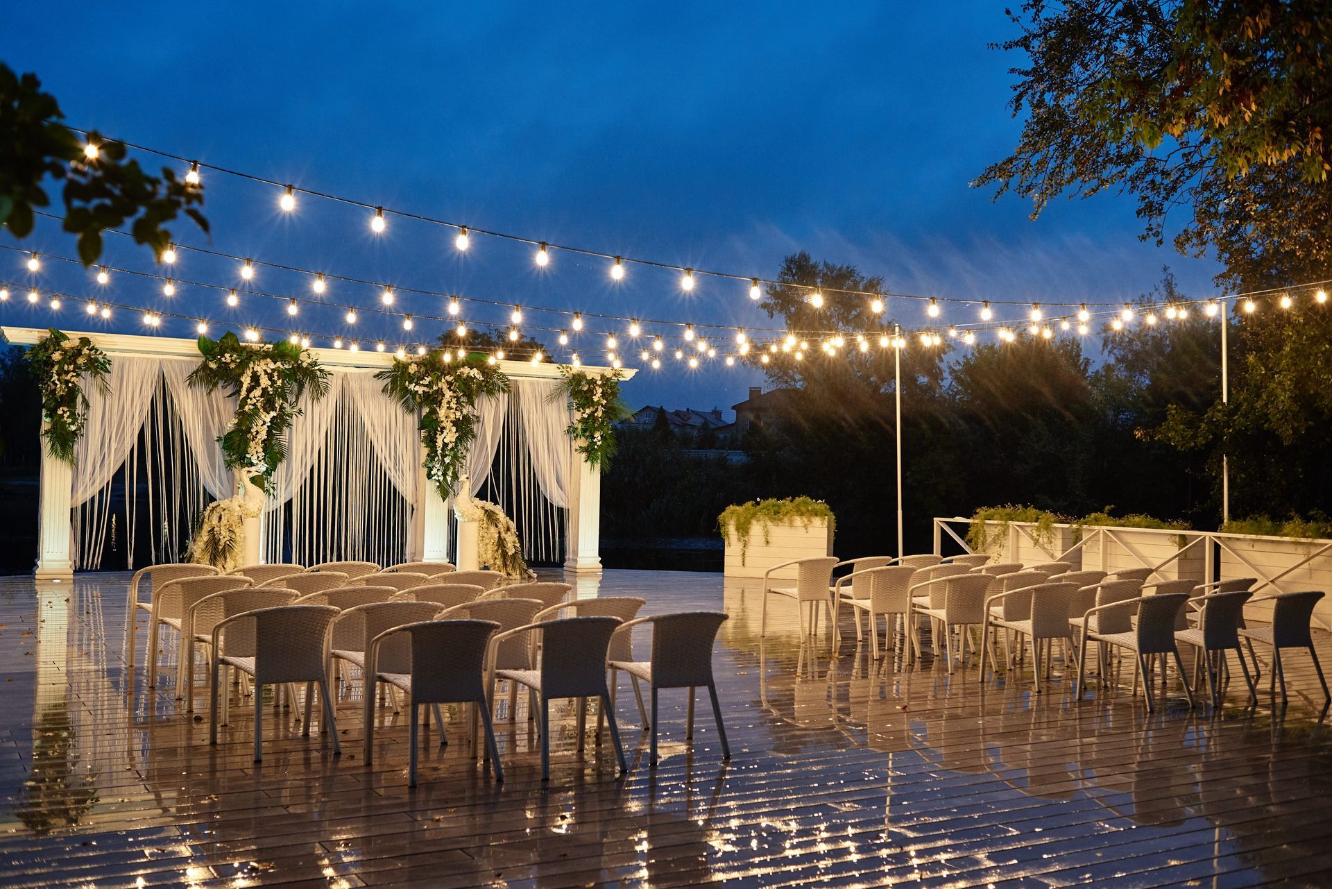 Outdoor evening wedding reception with string lights, white drapery, and rows of empty chairs by the water
