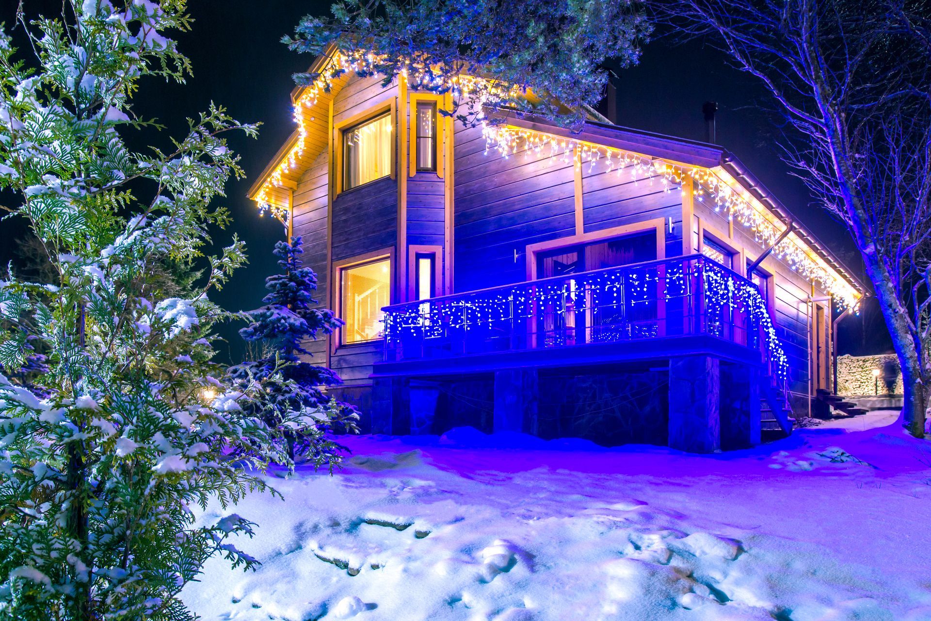 Snowy cabin at night glowing with blue and warm lights under trees