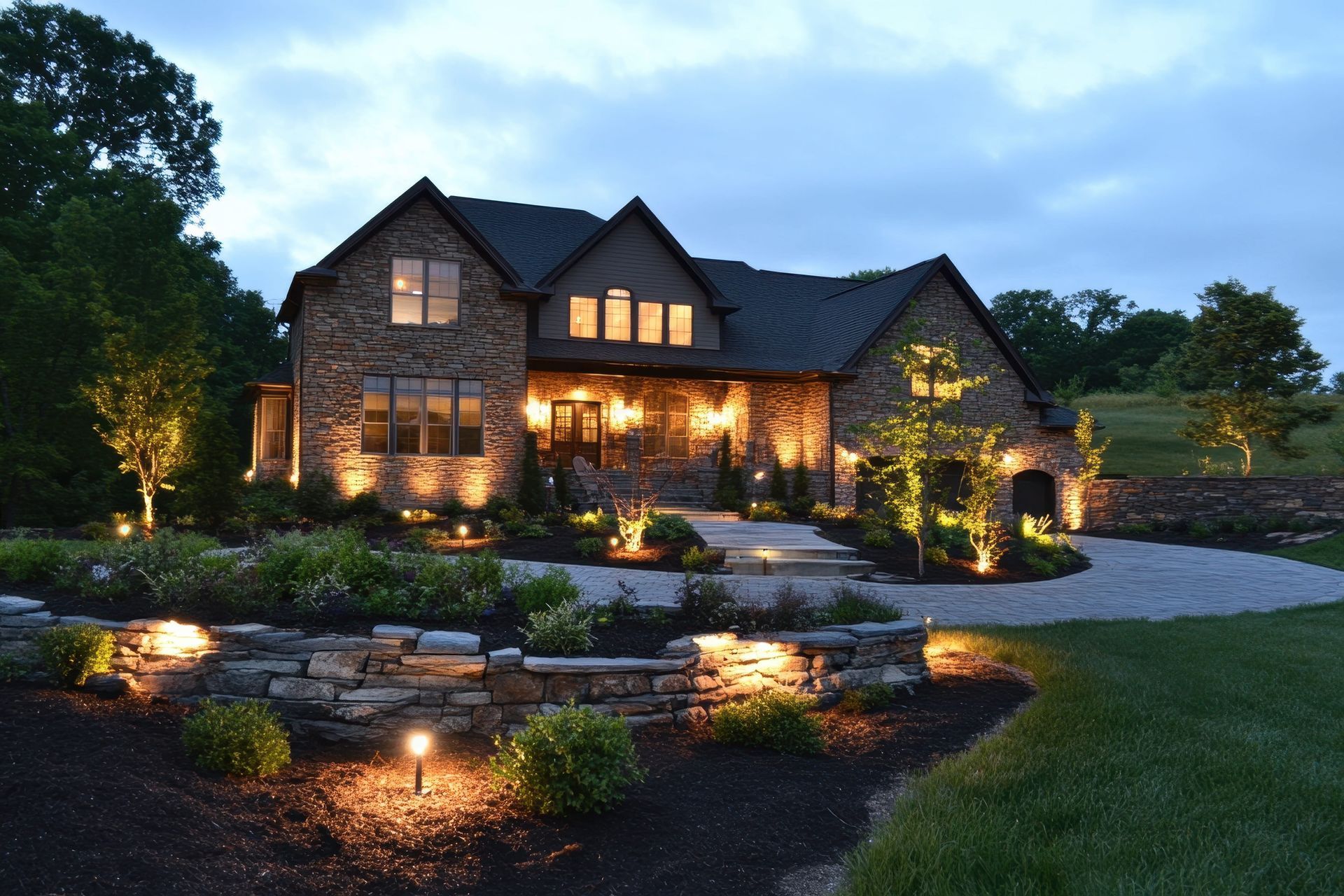 Large stone house at dusk with warm porch lights, landscaped garden, and curved driveway