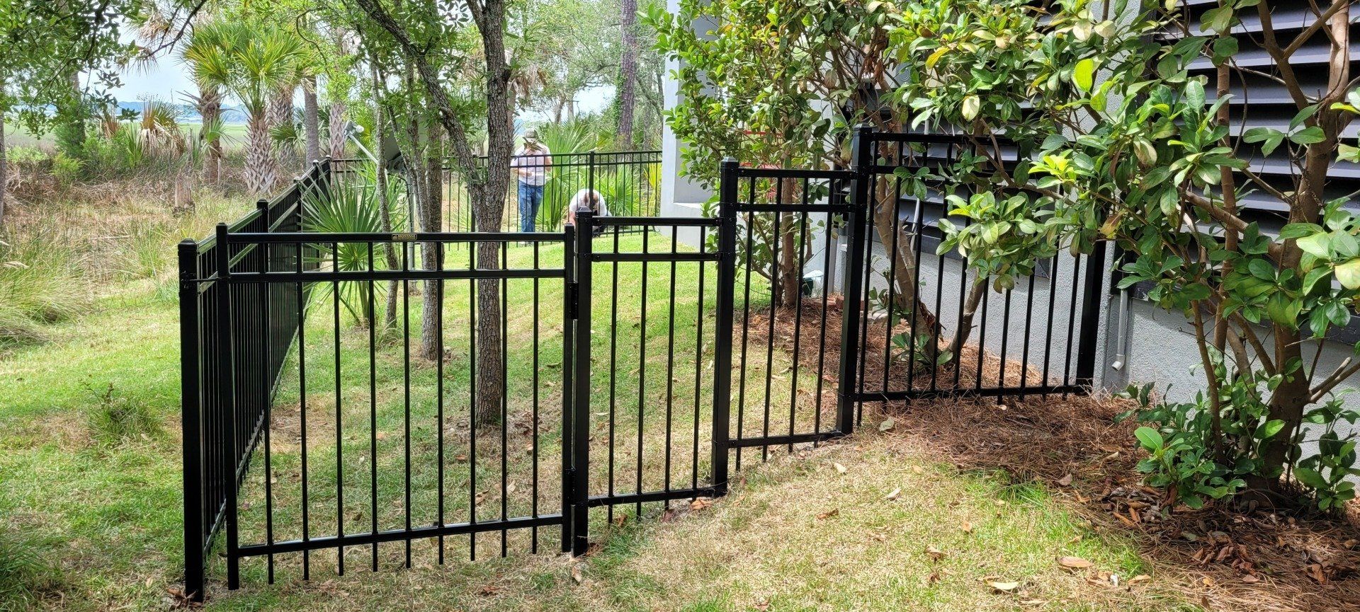A black metal fence with a gate is surrounded by grass and trees.