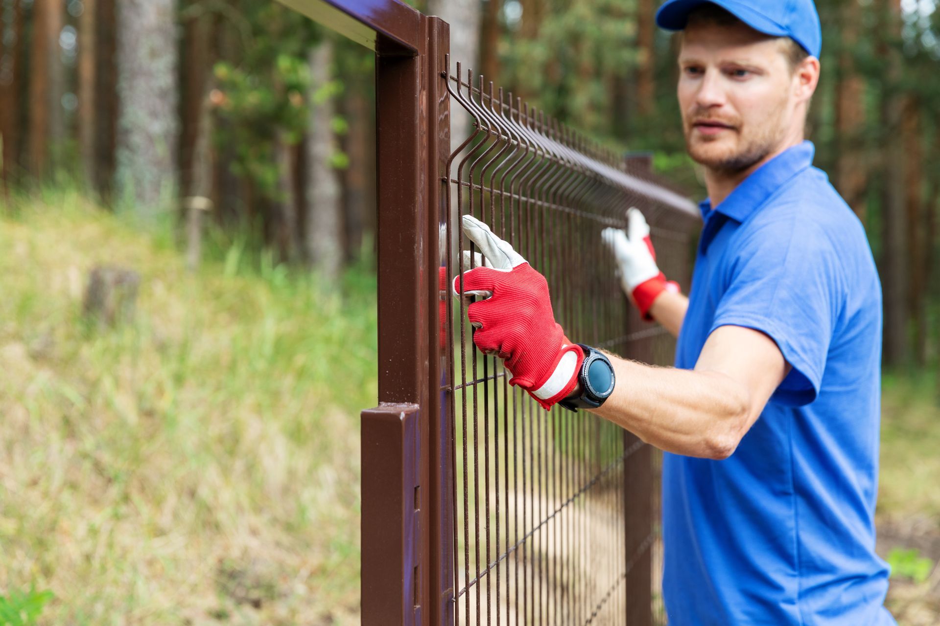 Worker in gloves installing a metal fence panel outdoors near a wooded area.
