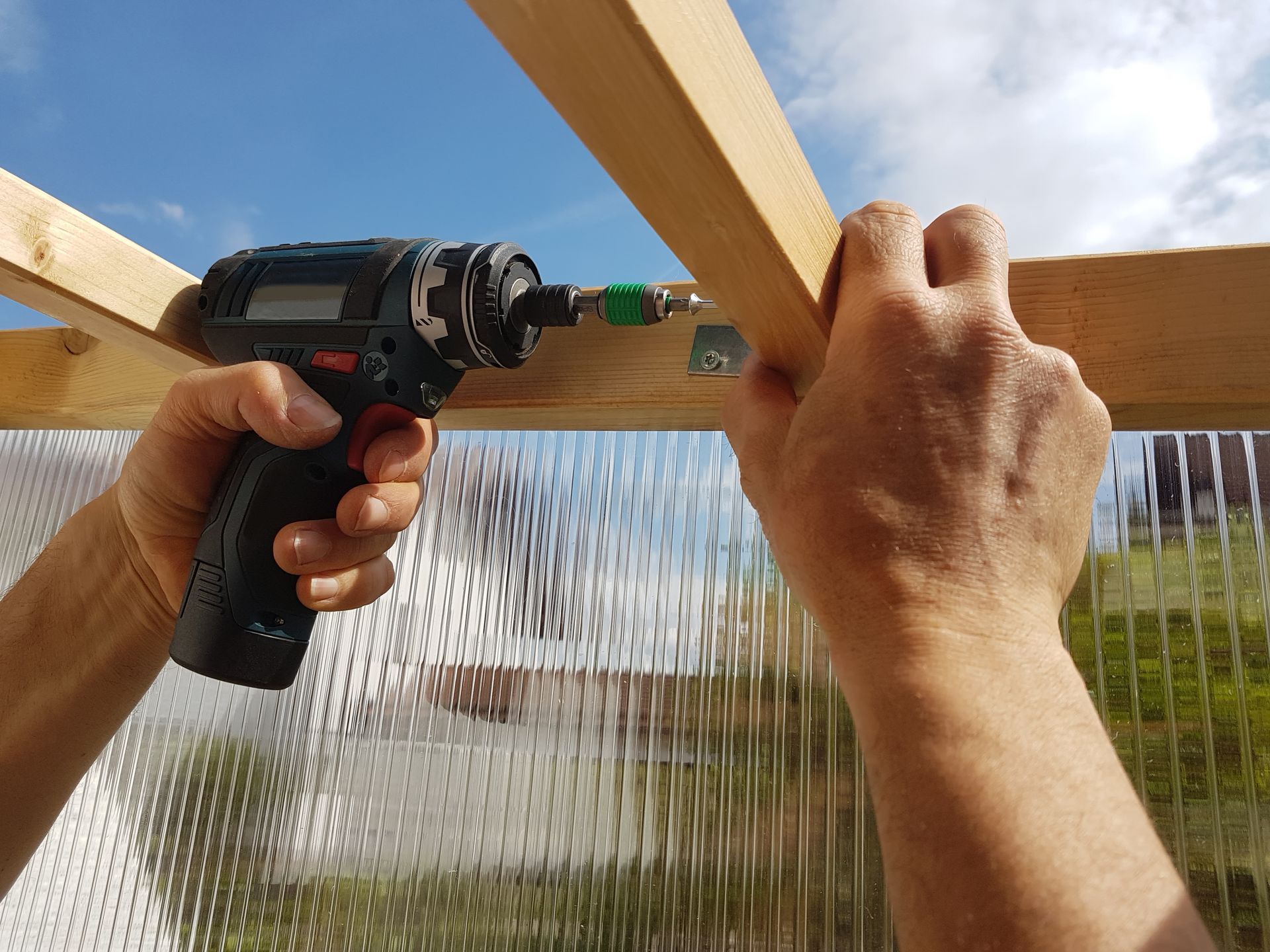 Hands using a drill to secure wooden beams, against a plastic panel backdrop, outdoors.