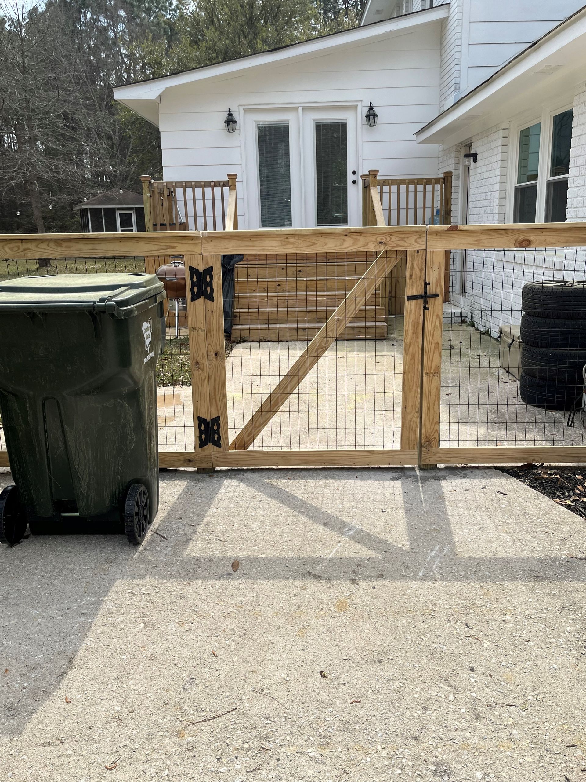 A green trash can is sitting next to a wooden gate in front of a house.