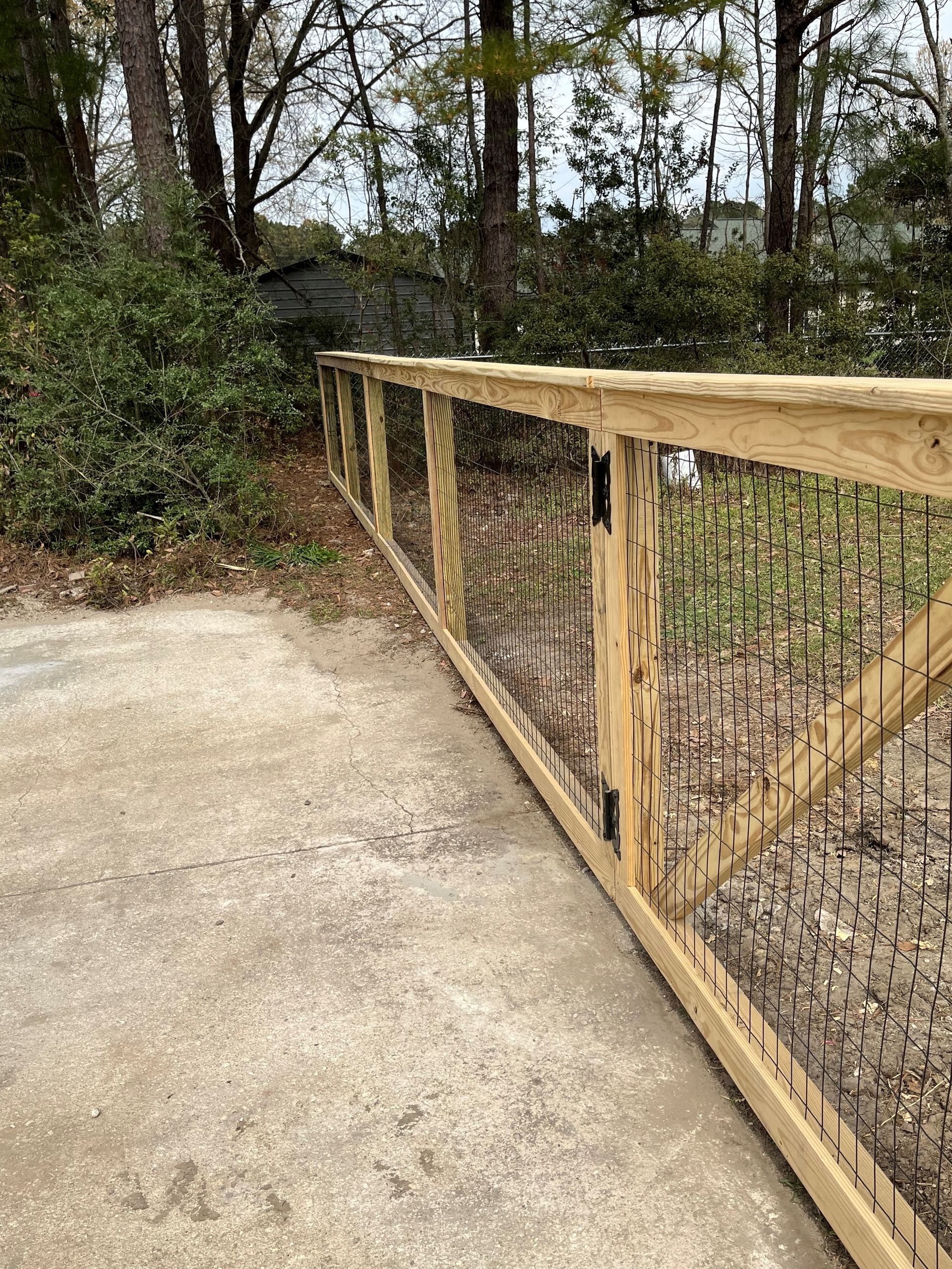 A wooden fence surrounds a concrete driveway with trees in the background.