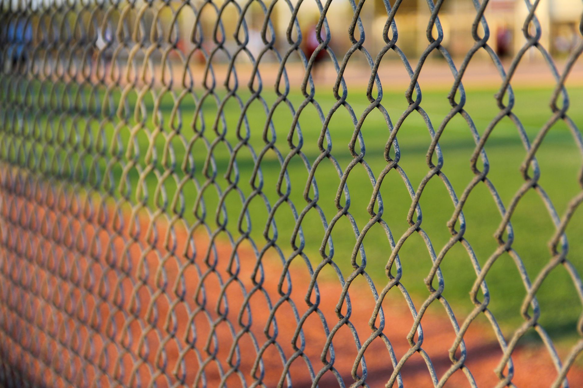 A close up of a chain link fence with a baseball field in the background.