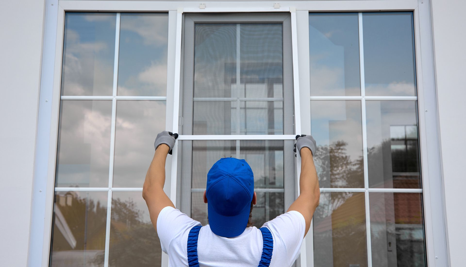 Person in blue overalls installing a screen window. Outdoors, daytime.