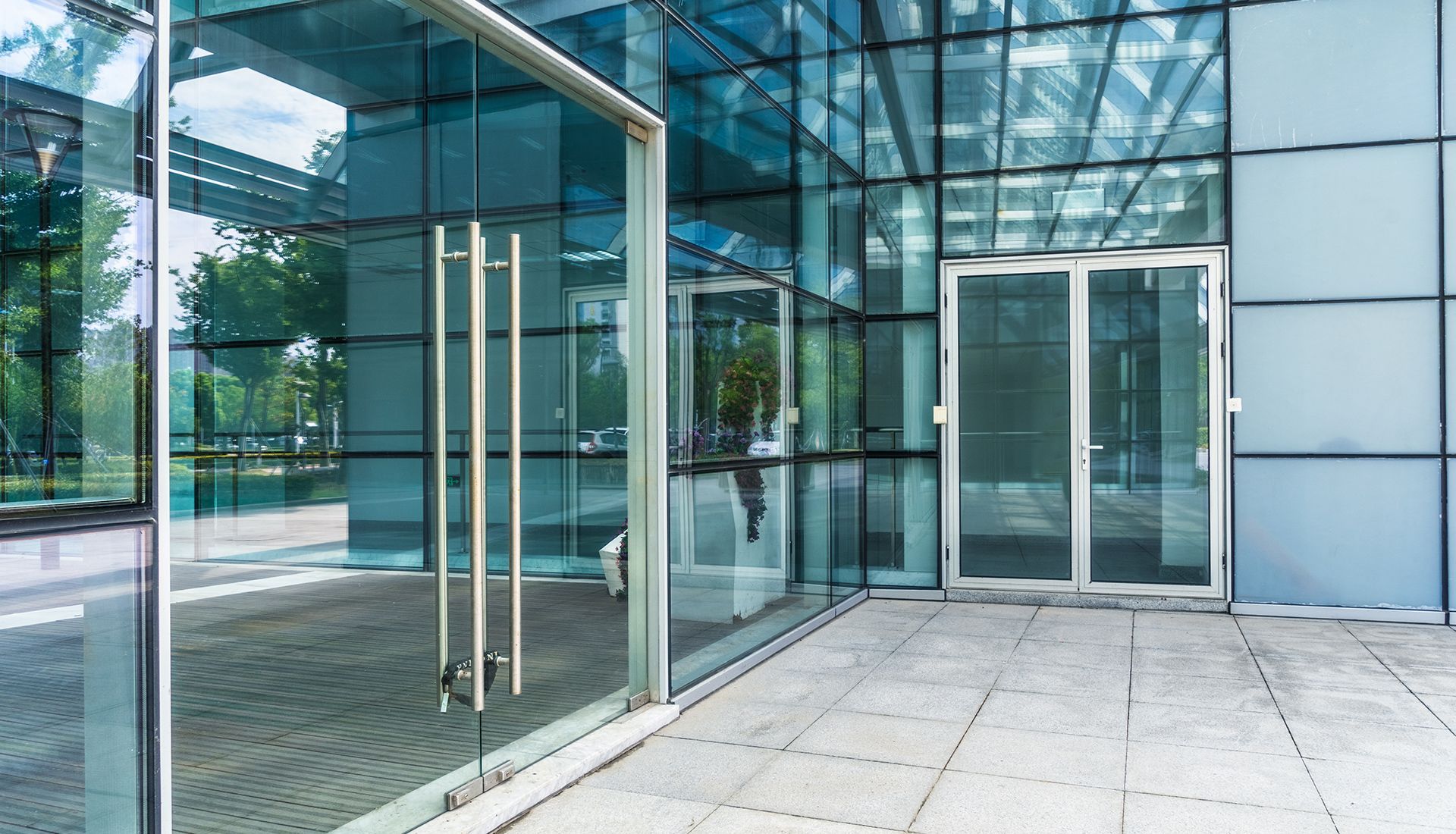 Modern glass building entrance with silver handles and doors, concrete walkway.