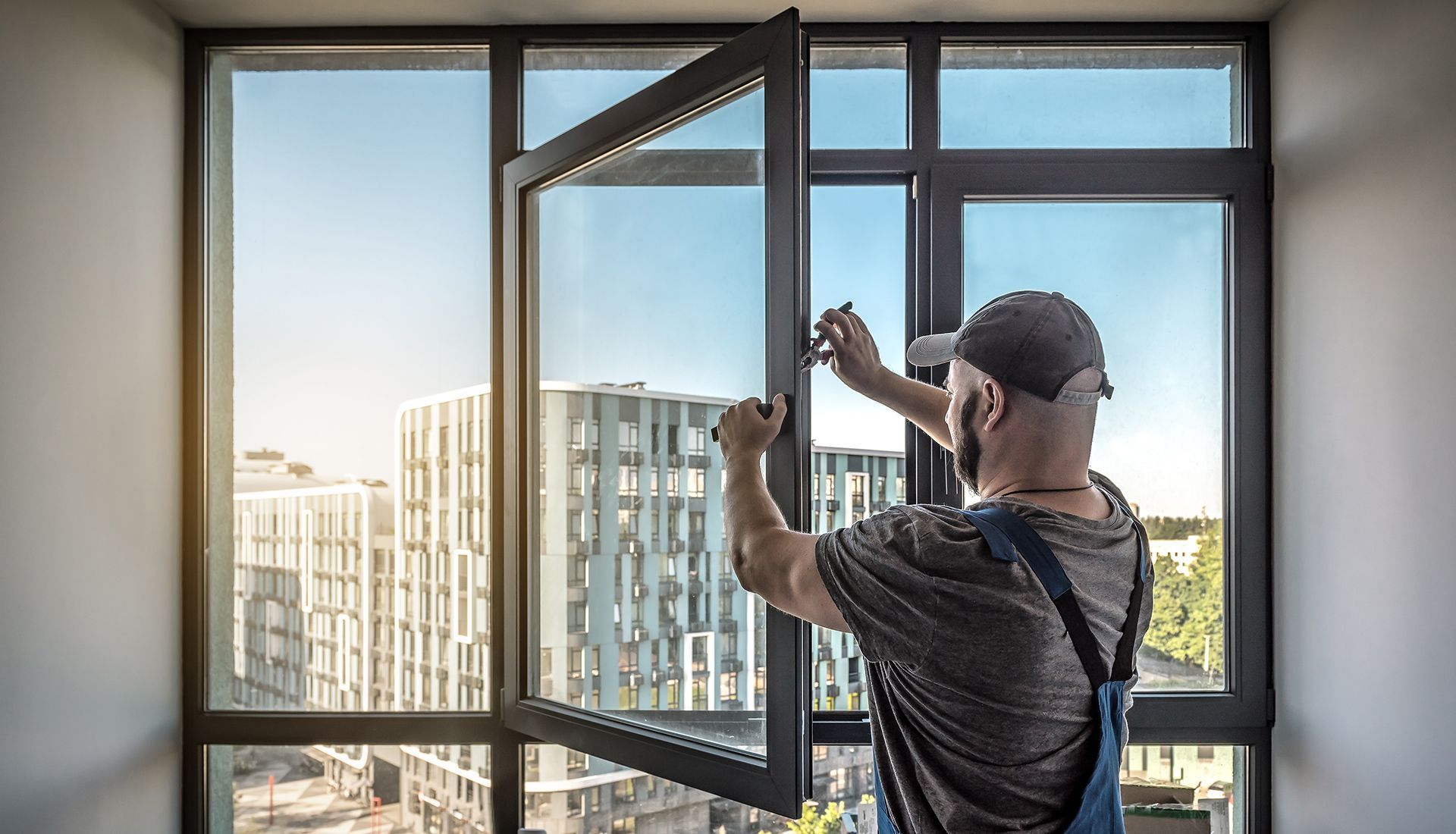 Man in overalls fixing an open, dark-framed window in a room with a city view.