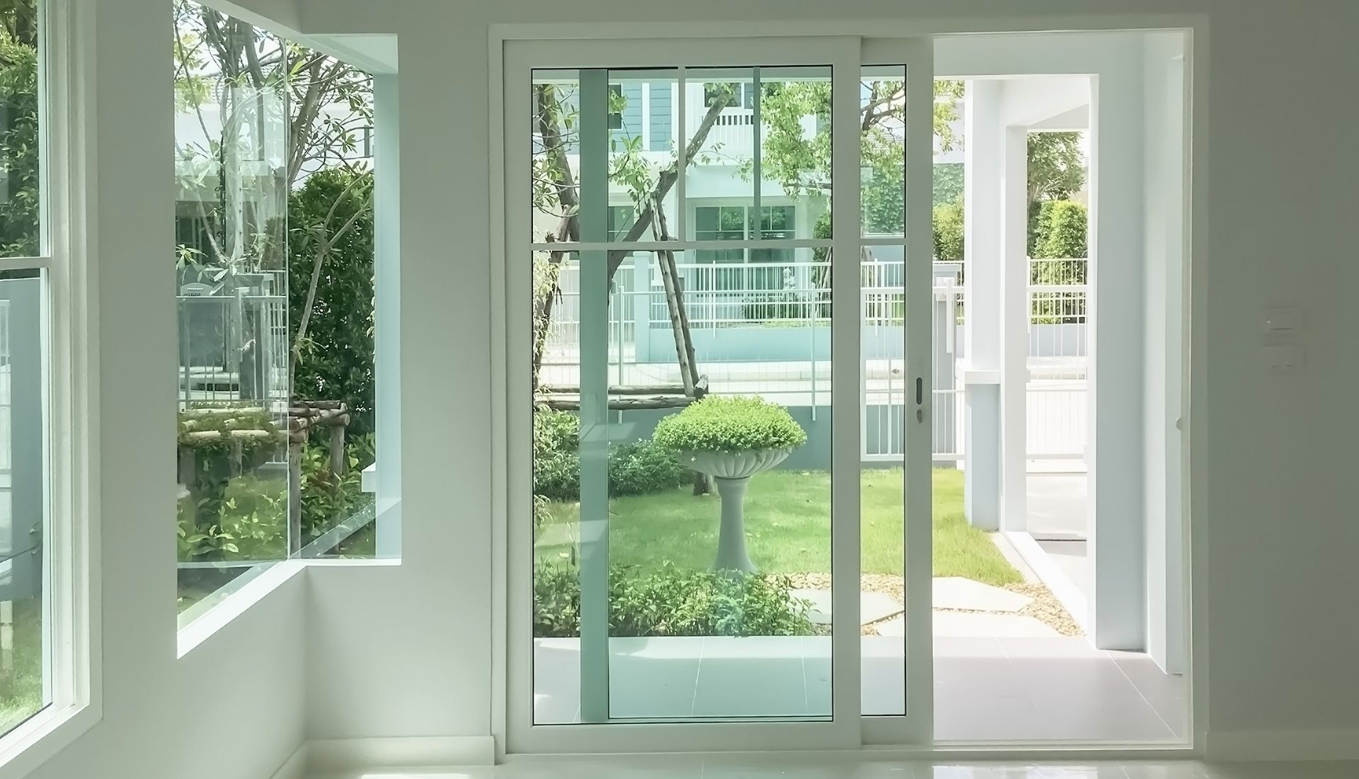 White-walled room with open sliding glass doors and windows, showcasing a garden and a second house.