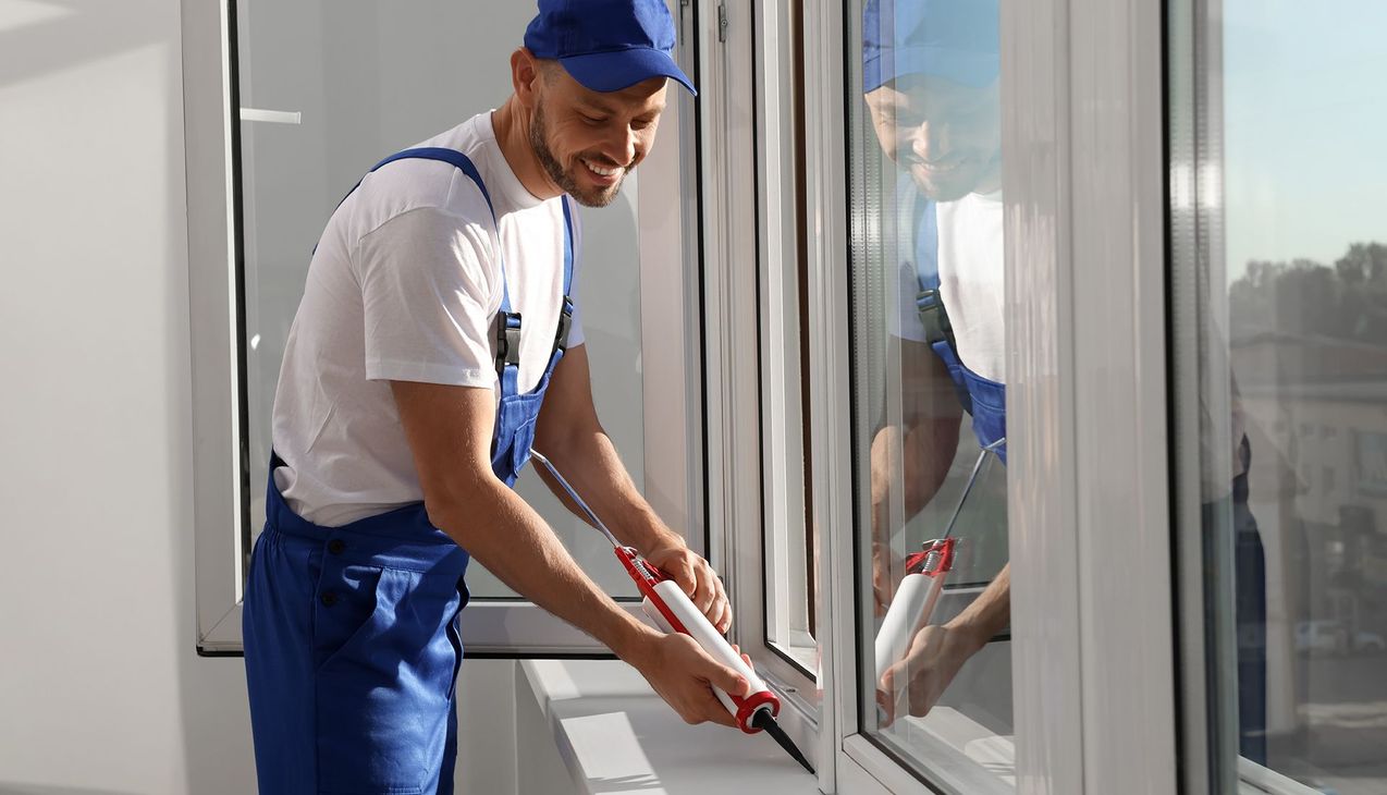 Man in blue overalls caulking a white window frame, smiling.