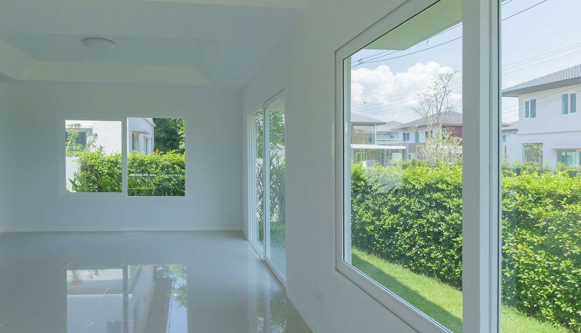 Empty white room with windows, green hedges and houses visible outside.