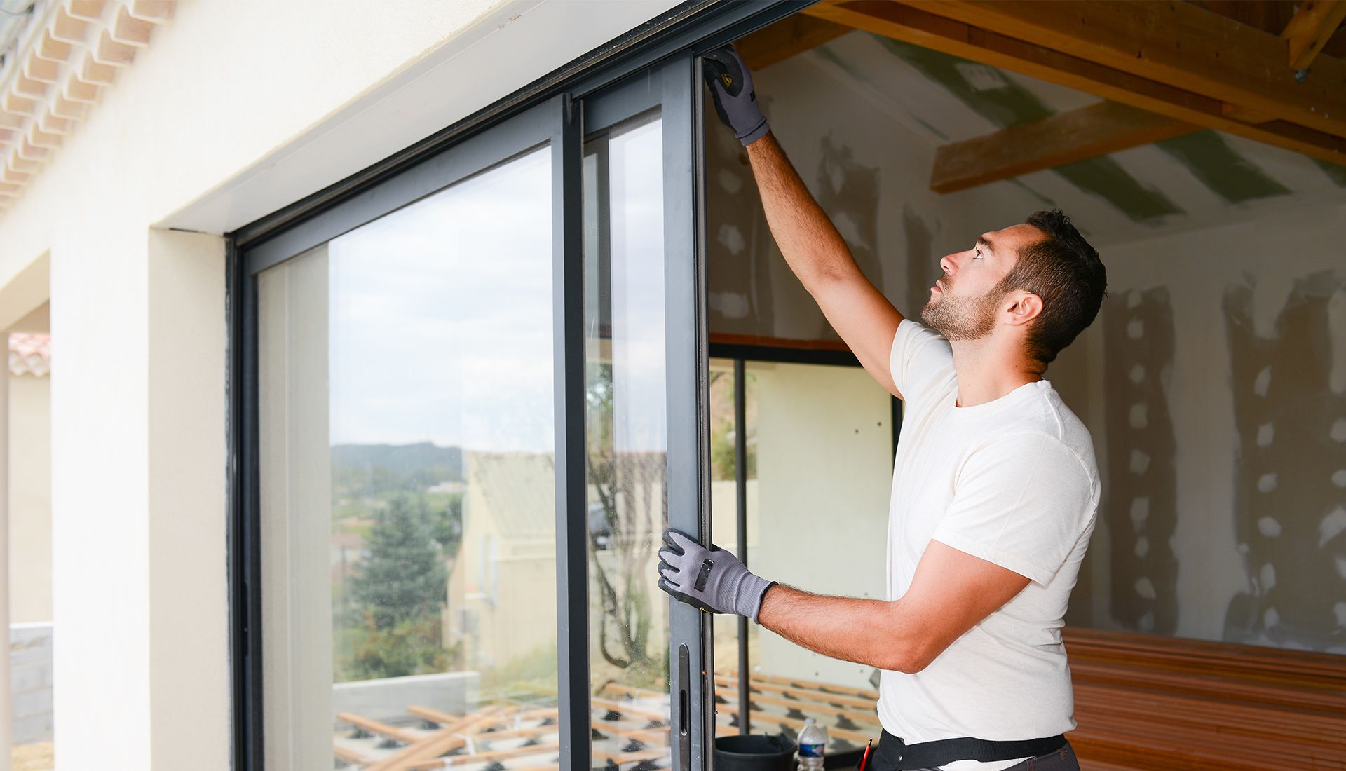 Man installing a sliding glass door; wearing gloves, reaching up, in a new construction setting.