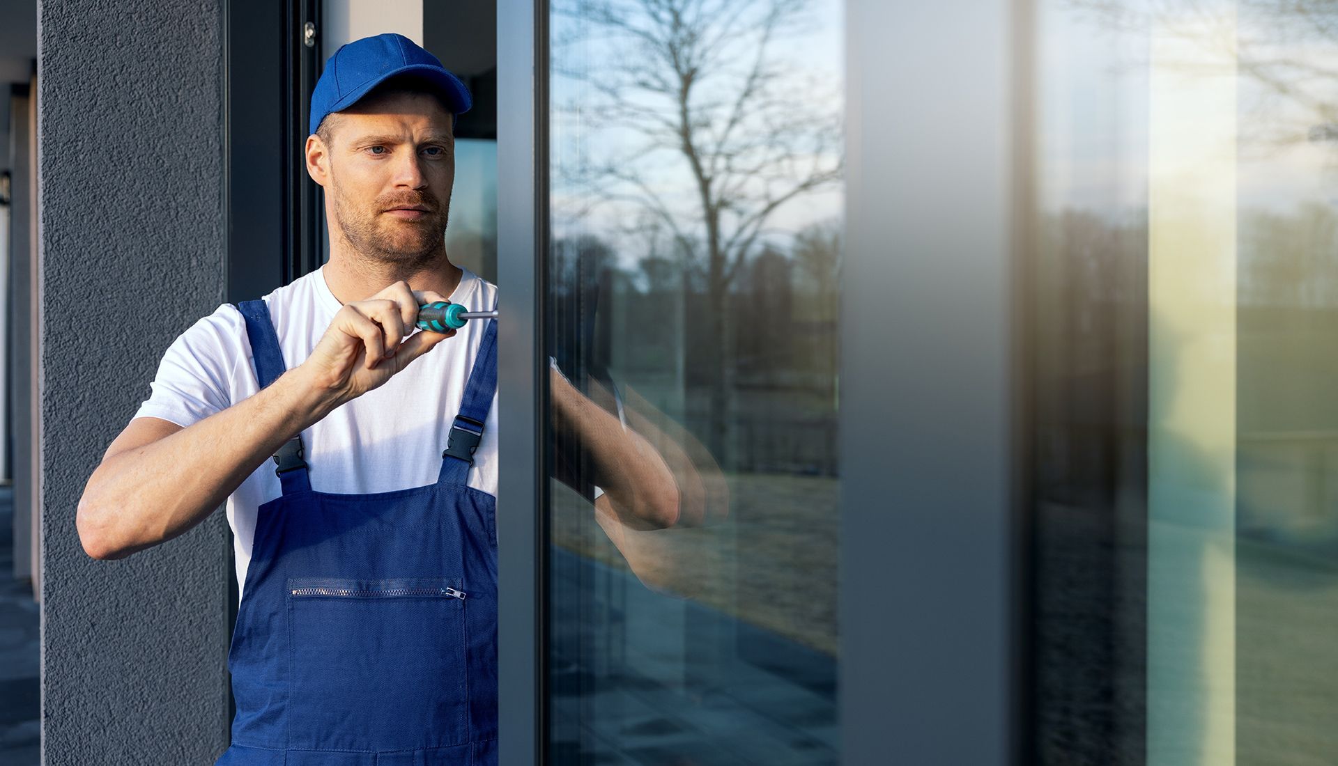 Man in blue overalls and cap, fixing a window with a screwdriver outdoors.