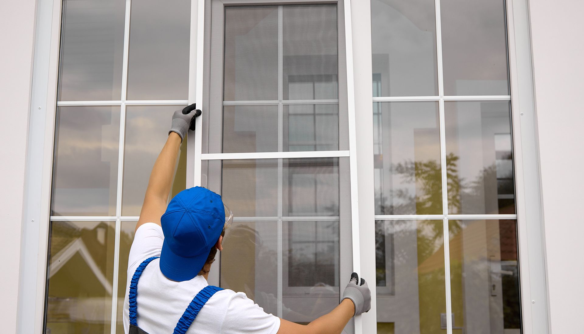 Man installing a white window screen, wearing gloves and a blue cap, outdoors.