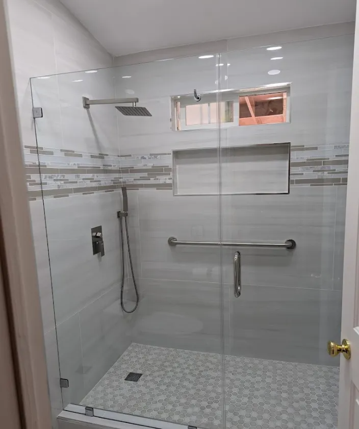 Glass-enclosed shower with white and beige tile. Features a handheld shower, grab bar, and built-in shelf.