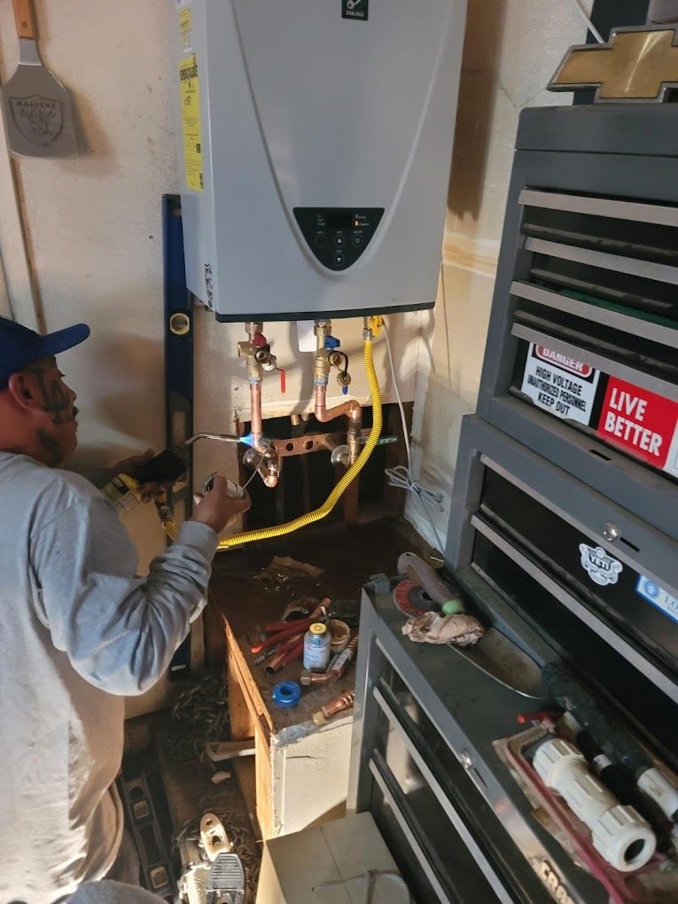 A technician installs pipes beneath a wall-mounted tankless water heater in a garage workspace.