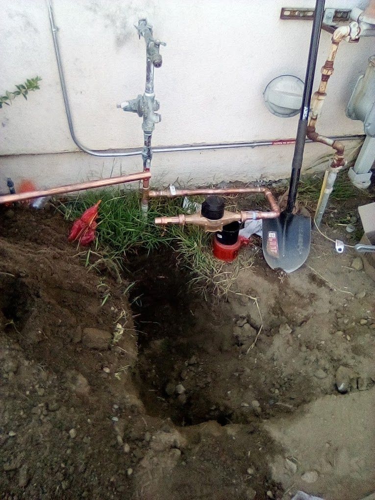 A copper plumbing repair on an outdoor wall near a trench with a shovel and a black valve.