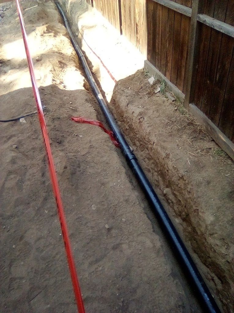 A black pipe lies in a narrow dirt trench alongside a wooden fence, with a red utility marking line running parallel.
