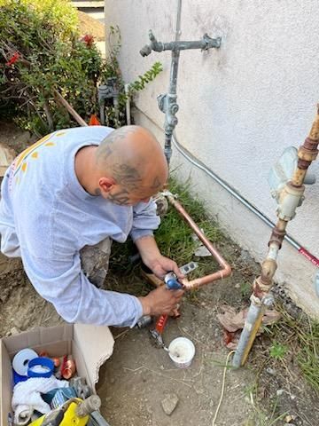 A person kneels by a house foundation, soldering copper pipes near a water meter setup.