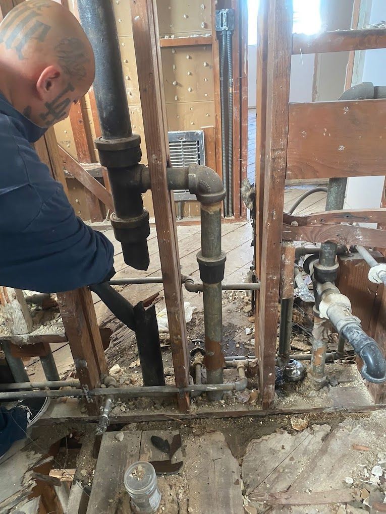 A person works on exposed plumbing pipes inside the wooden wall frame of a room under construction.