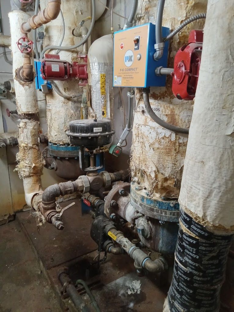 Industrial mechanical equipment, including insulated pipes, valves, and a control box, in a utility room.