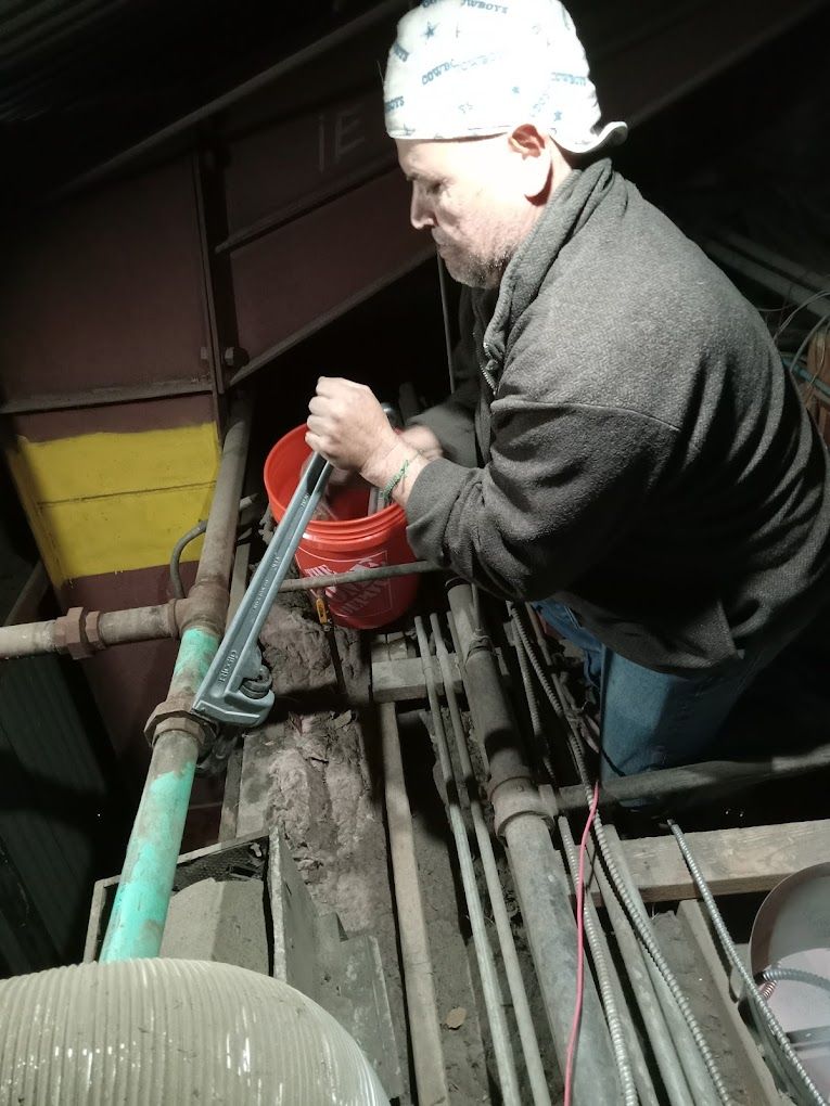 A person wearing a white cap uses a pipe wrench on a green industrial pipe, with a red bucket nearby in a dark setting.