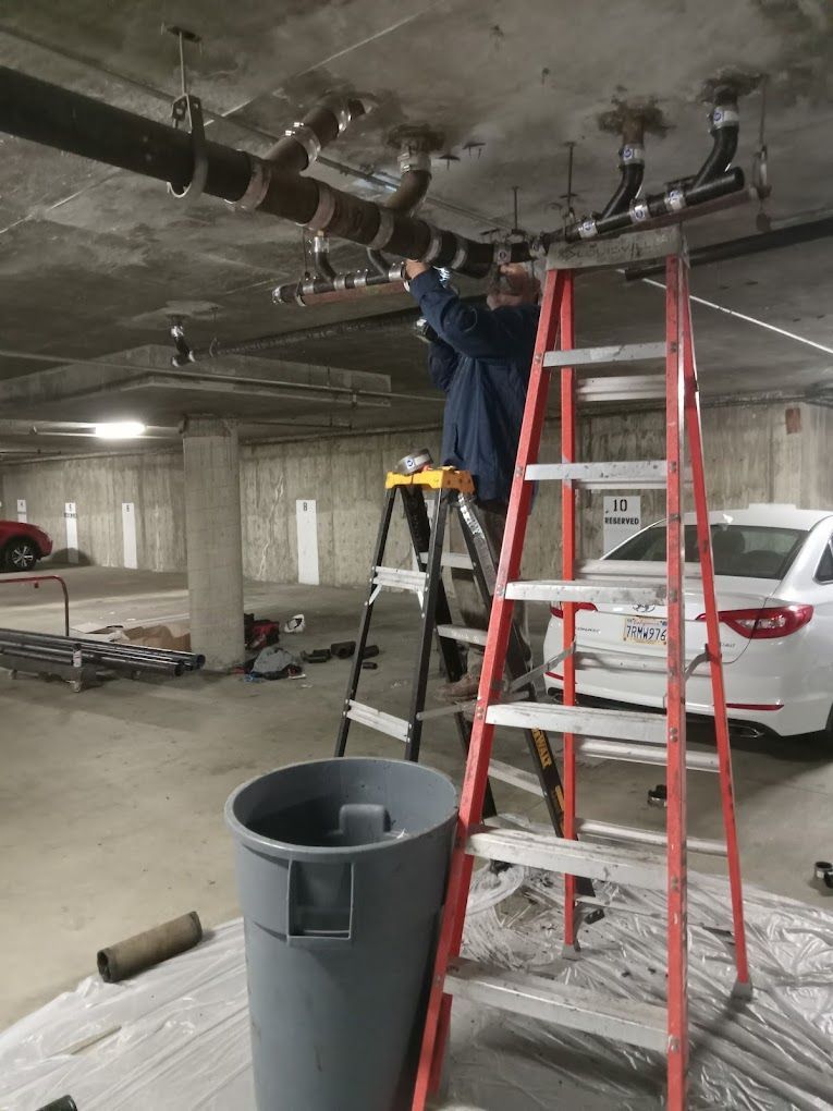 A person stands on a ladder working on pipes attached to a concrete ceiling in a parking garage.